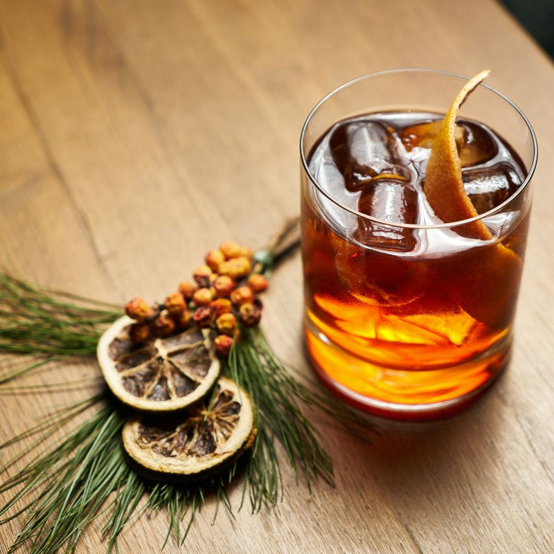 Birch Old Fashioned in a clear glass with needles and dried oranges laying on the wood table next to the glass.