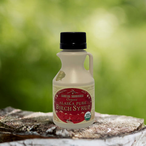 Kahiltna Birchworks Organic Alaska Pure Birch Syrup, 3.4 fl oz plastic jug on a birch log with a green outdoor background