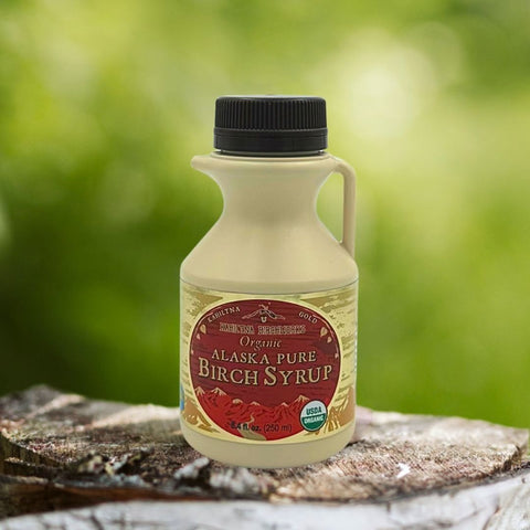 Kahiltna Birchworks Organic Alaska Pure Birch Syrup, 8.4 fl oz plastic jug on a birch log with a green outdoor background