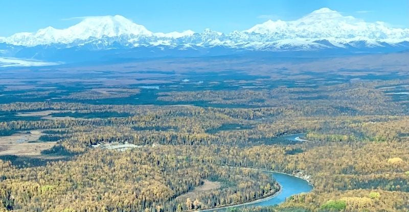 Aerial view of a Alaska river winding through a forest with snow-capped mountains in the background.