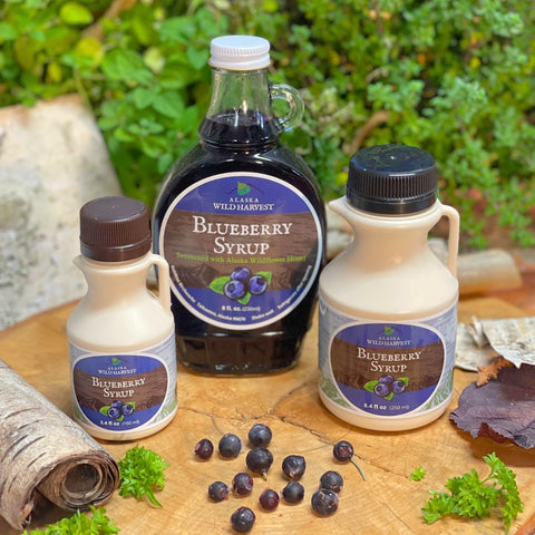Three bottles of Blueberry Syrup from Alaska Wild Harvest on a wooden surface with blueberries and greenery.