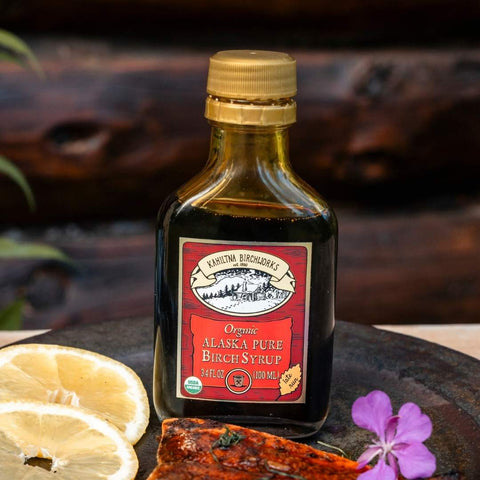 A bottle of Kahiltna Birchworks Organic Alaska Pure Birch Syrup, 4 fl oz, displayed on a dark rustic surface with lemon slices, a piece of salmon, and a purple flower.