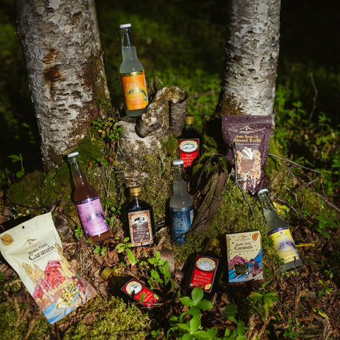 A collection of artisanal beverages and packaged snacks from Kahiltna Birchworks and Hammers Family Birch arranged on the forest floor between two birch trees. The assortment includes bottles and bags of drinks and sweets with colorful labels, set in a mossy, outdoor woodland environment.