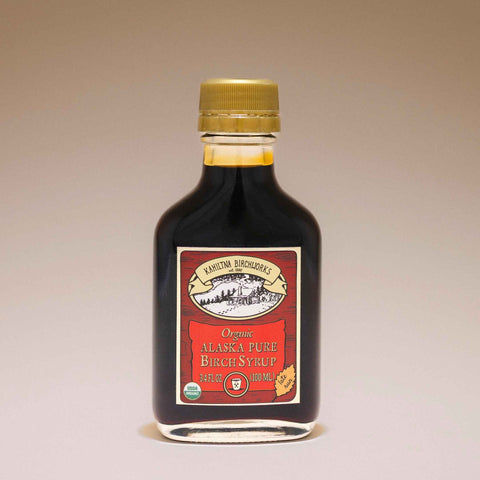A bottle of Kahiltna Birchworks Organic Alaska Pure Birch Syrup, featuring a dark brown liquid, a gold cap, and a red and beige label, set against a neutral beige background.