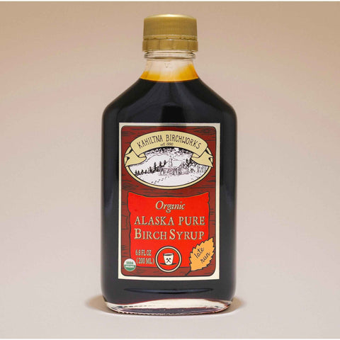 A bottle of Kahiltna Birchworks Organic Alaska Pure Birch Syrup with a gold cap and red label, set against a plain beige background.