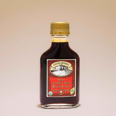 A bottle of Kahiltna Birchworks Organic Alaska Pure Birch Syrup with a gold cap and a red and cream label, set against a plain beige background.