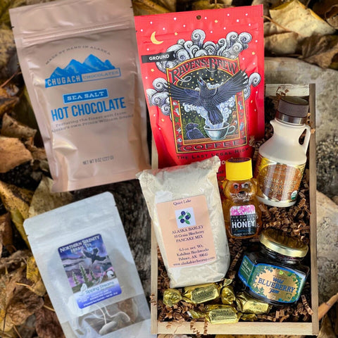 The Northern Sunrise Gift Crate. Assorted food items, including hot chocolate, coffee, honey, and herbal tea in a wooden box on a leafy background.