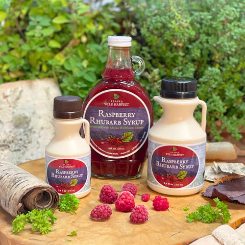 Three bottles of raspberry rhubarb syrup on a wooden board with fresh raspberries and leaves.