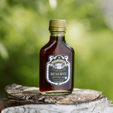 3.4 oz bottle of Reserve pure birch syrup on a wooden stump with a blurred green background