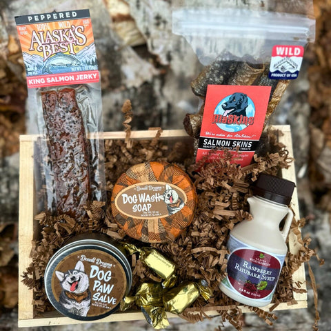 Collection of pet supplies including soap, paw salve, and salmon skins as well as king salmon jerky and raspberry rhubarb syrup for the pet owner in a gift crate
