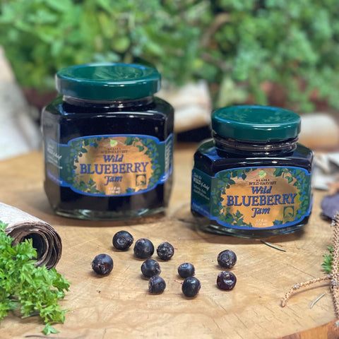 Two jars of Wild Blueberry Jam on a wooden surface with blueberries around, greenery in the background.