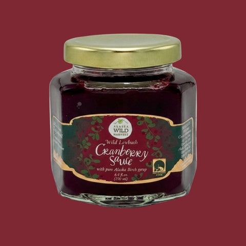 A glass jar of Alaska Wild Cranberry Sauce with a gold lid and a dark green label, centered against a solid dark red background.