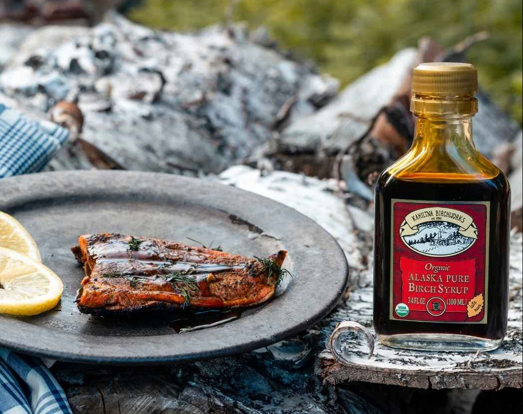 A bottle of Kahiltna Birchworks Organic Alaska Pure Birch Syrup stands on a rustic log beside a plate of grilled salmon with lemon slices, set outdoors near a campfire.