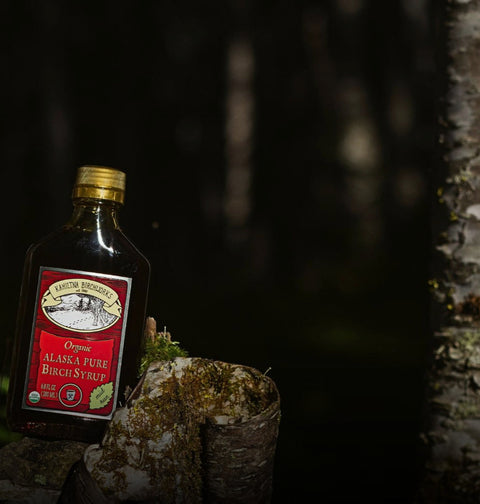 A bottle of Alaska Pure Birch Syrup by Kahiltna Birchworks of Alaska, styled on a mossy log in a dark, forest-like setting with birch tree bark on the right.