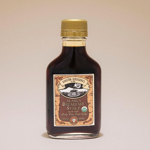 A bottle of Kahiltna Birchworks Alaska Breakfast Syrup with a gold cap and a brown and beige label, set against a plain beige background.
