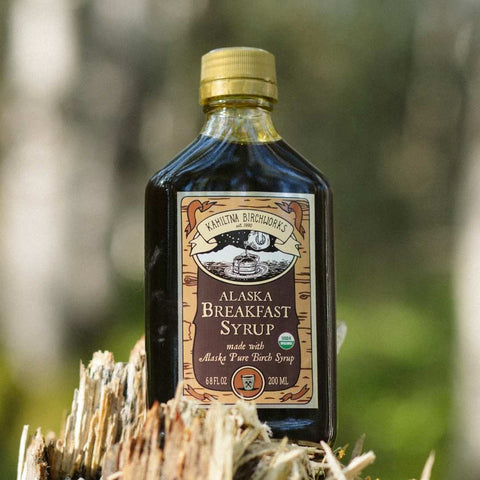 A bottle of Kahiltna Birchworks Alaska Breakfast Syrup stands on a piece of weathered wood with a blurred outdoor forest background.