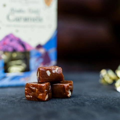 Three pieces of birch caramel candy with visible nuts stacked on a dark surface, with a blurred package of caramel candies in the background.