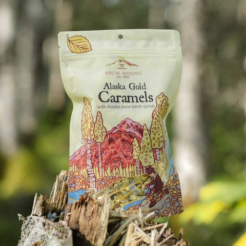 A stand-up pouch of Kahiltna Birchworks Alaska Gold Caramels with a colorful forest illustration, photographed outdoors on a tree stump with a blurred forest background.