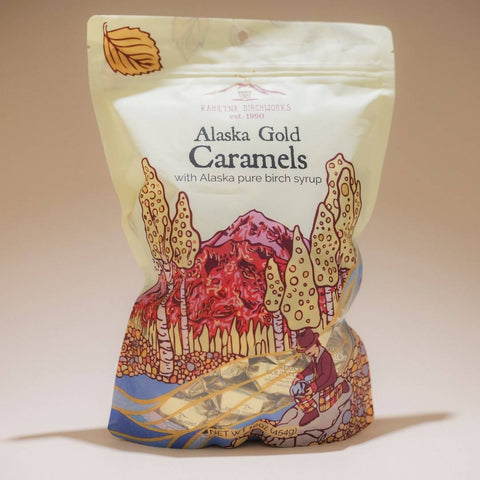 A stand-up pouch of Kahiltna Birchworks Alaska Gold Caramels, featuring a cream-colored background with an illustrated graphic of a mountain, birch trees, and a child in the lower half.