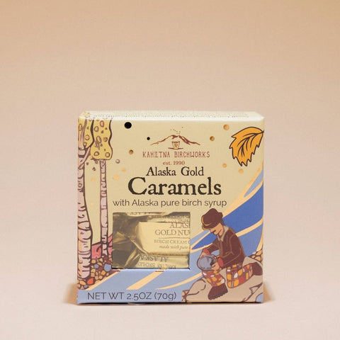 A boxed pack of Alaska Gold Caramels with Alaska pure birch syrup, shown against a solid beige background. The packaging features a cream-colored background with illustrations of a birch tree, an autumn leaf, and a Native American figure, along with a clear window displaying the wrapped caramels inside.