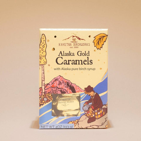 A package of Kahiltna Birchworks Alaska Gold Caramels, featuring a cream-colored box with colorful illustrations of a mountain, a woman making caramel, and a gold foil window showing the wrapped caramels inside.