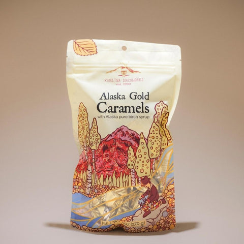 A stand-up pouch of Khetna Barcholars Alaska Gold Caramels, featuring a cream-colored background with an illustrated landscape of red mountains, golden trees, and a river.