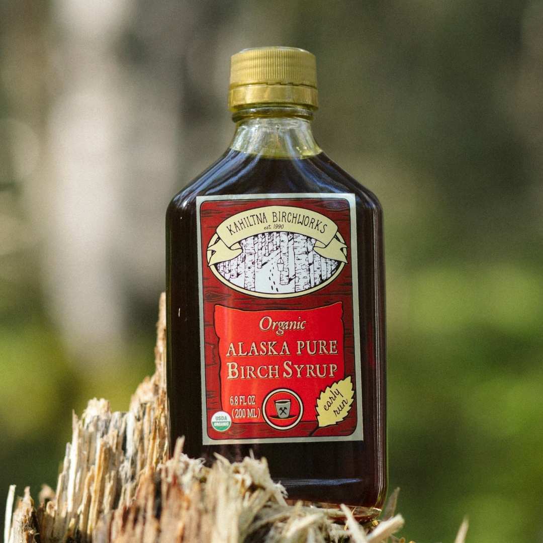 A bottle of Kahiltna Birchworks Organic Alaska Pure Birch Syrup with a gold cap, set on a weathered tree stump with a blurred forest background.
