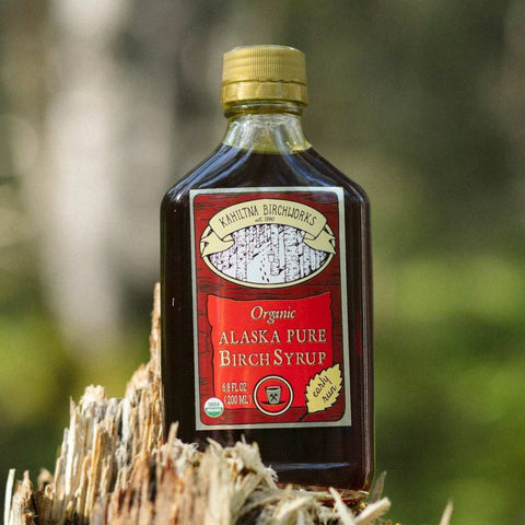 A bottle of Kahiltna Birchworks Organic Alaska Pure Birch Syrup with a gold cap, set on a weathered tree stump with a blurred forest background.