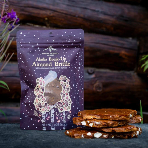 A stand-up pouch of Kahiltna Birchworks Alaska Break-Up Almond Brittle stands on a dark surface with a stack of brittle pieces in front. The pouch is dark purple with white text and an illustrated bear made of almonds.