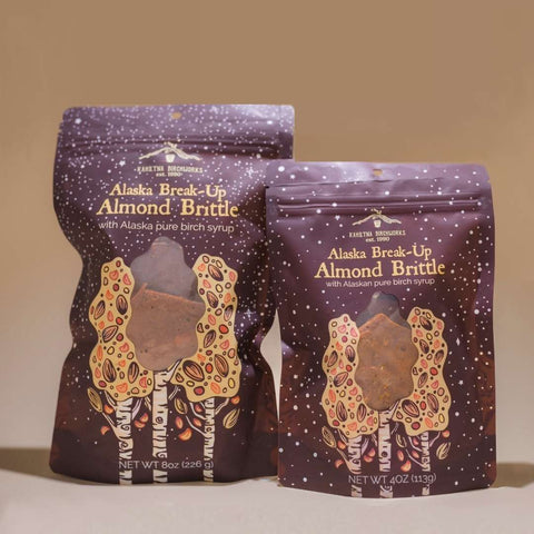 Two stand-up pouches of Kahiltna Birchworks Alaska Break-Up Almond Brittle with Alaska pure birch syrup, shown on a warm beige studio background. The packaging is dark purple with white speckles, gold lettering, and an illustration of almond trees with almonds.