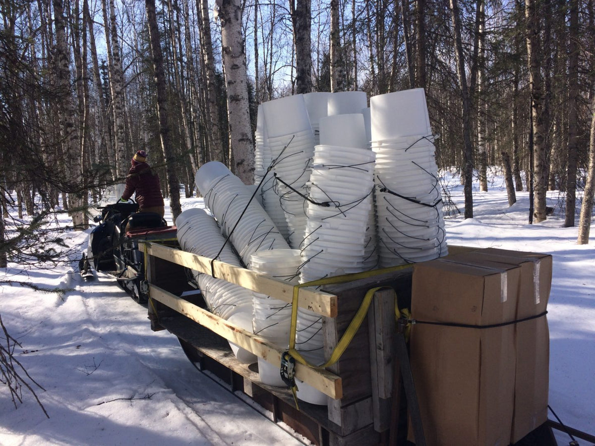 Wooden trailer loaded with large white plastic birch sap collection bins in a snowy forest.