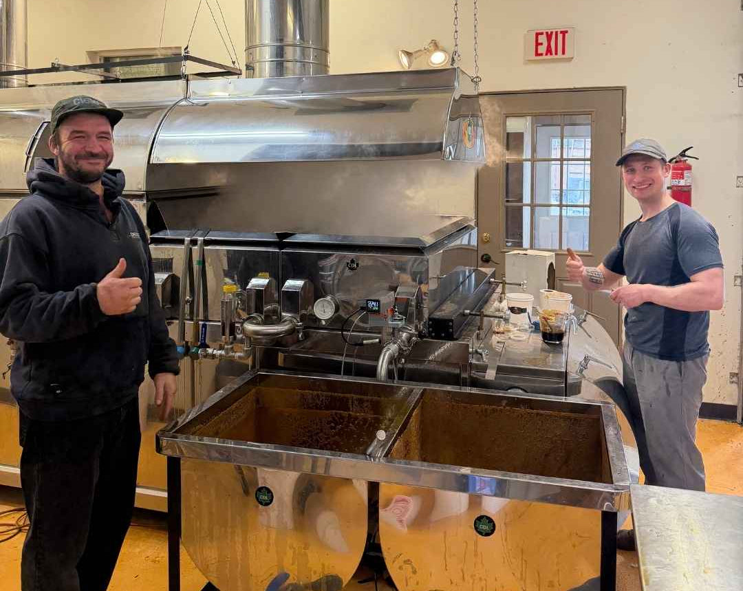 Two men standing on either side of a large stainless steel birch sap distillation machine in an indoor workshop