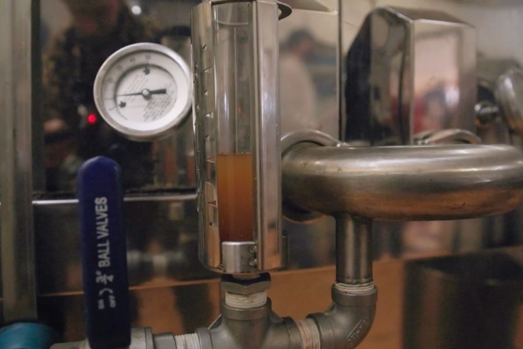 A close-up of a stainless steel liquid transfer setup for cooking birch syrup with a clear vertical tube showing amber liquid, a round pressure gauge, and a blue ball valve labeled 'BALL VALVES'.