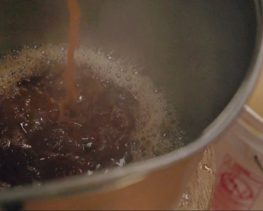 A close-up shot of a dark brown liquid that is birch sap in the process of being made into birch syrup being poured into a metal pot with active foam and bubbles on the surface.