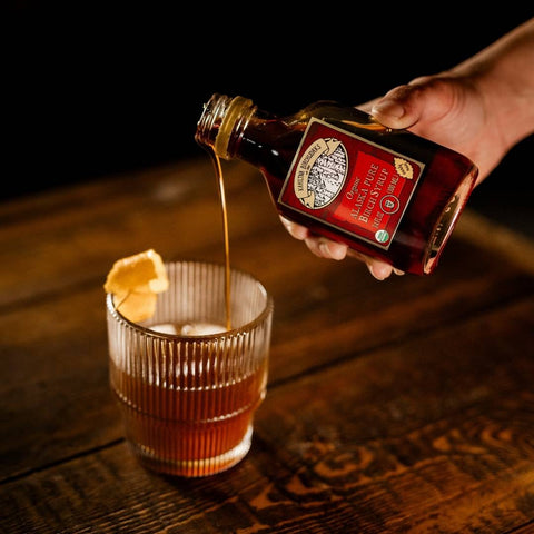 A hand pours birch syrup from a small bottle labeled 'Organic Alaska Pure Birch Syrup' into a ribbed glass tumbler on a dark wooden table.