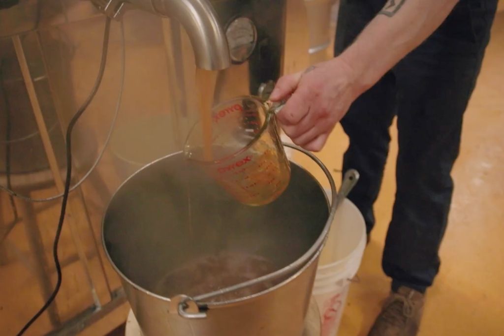 A person pouring liquid birch syrup from a measuring cup into a large metal bucket in a birch syrup workshop setting.
