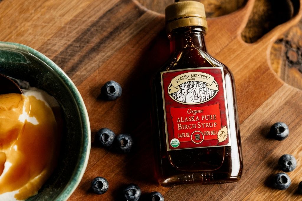 A bottle of Kahiltna Birchworks Organic Alaska Pure Birch Syrup placed on a wooden cutting board next to a bowl of whipped cream with a drizzle of syrup, with scattered blueberries around it.