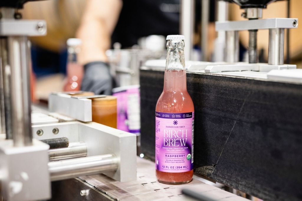 A bottle of Raspberry Birch Brew, a fermented birch water sparkling beverage with a purple label stands on a conveyor belt in a production setting, with machinery and a blurred worker in the background.