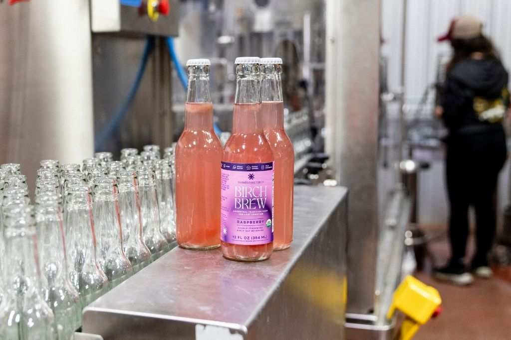 Two glass bottles of raspberry fermenter birch brew, a birch sap beverage, with purple labels on a stainless steel conveyor belt in a factory setting, with empty glass bottles lined up on the left and a worker blurred in the background.