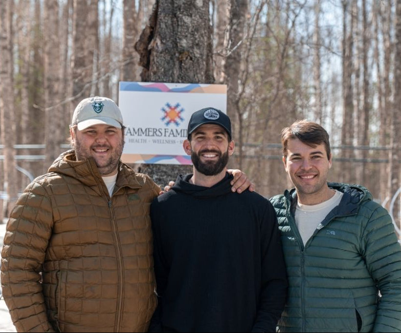 Three men standing outdoors in front of bare trees, with a sign reading 'Hammers Family Birch' behind them. The men are wearing winter jackets and caps, and one man has his arm around the others.
