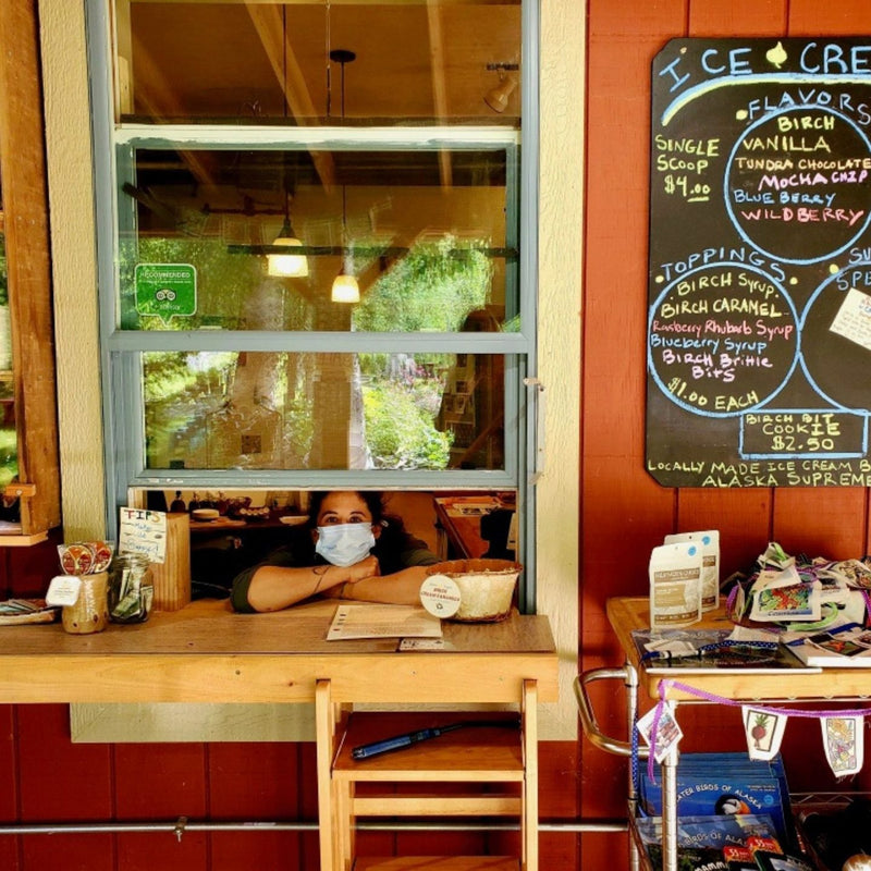Person wearing a face mask sitting at a counter in a cafe with an ice cream menu on the wall at the Kahiltna Birchworks shop.