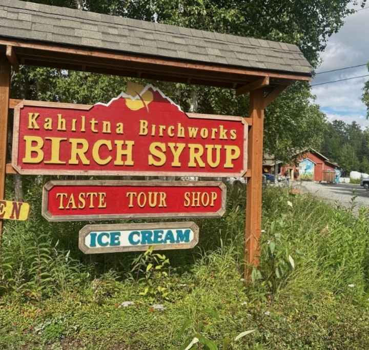 Sign for Kahlitna Birchworks Birch Syrup, Taste Tour Shop, and Ice Cream on a wooden post with trees and road in the background.