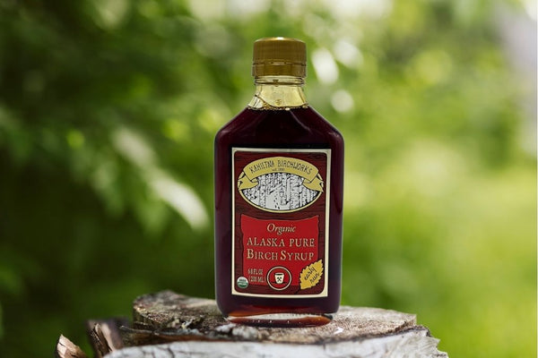A bottle of Kahiltna Birchworks Organic Alaska Pure Birch Syrup standing on a tree stump with a blurred green forest background.