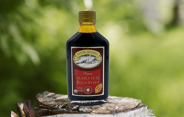 A bottle of Kahiltna Birchworks Organic Alaska Pure Birch Syrup standing on a cut tree stump with a blurred green outdoor background.