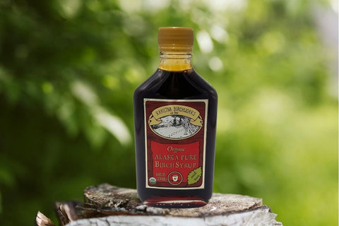 A bottle of Kahiltna Birchworks  Organic Alaska Pure Birch Syrup standing on a tree stump with a blurred green outdoor background.