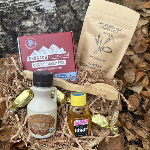 Assorted Alaska-themed products in the Sourdough and Sweet Gift Basket, including chocolate, sourdough starter, and honey on a wooden surface.