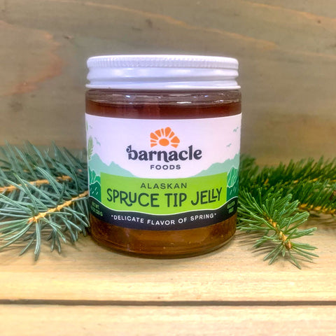 Jar of Alaskan Spruce Tip Jelly from Barnacle Foods with spruce branches on a wooden surface.