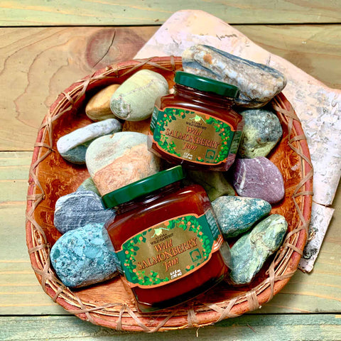 Two jars of wild salmonberry jam with colorful stones on a woven tray on a wooden surface.
